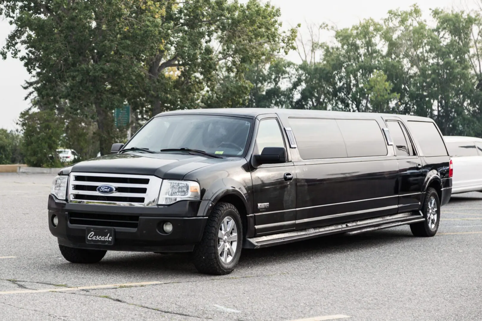 Image shows a black Ford Expedition stretch limousine parked on a road with green tress in the background.