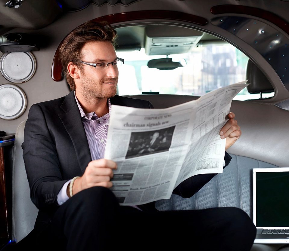 A man sitting in the back of a car reading a newspaper.